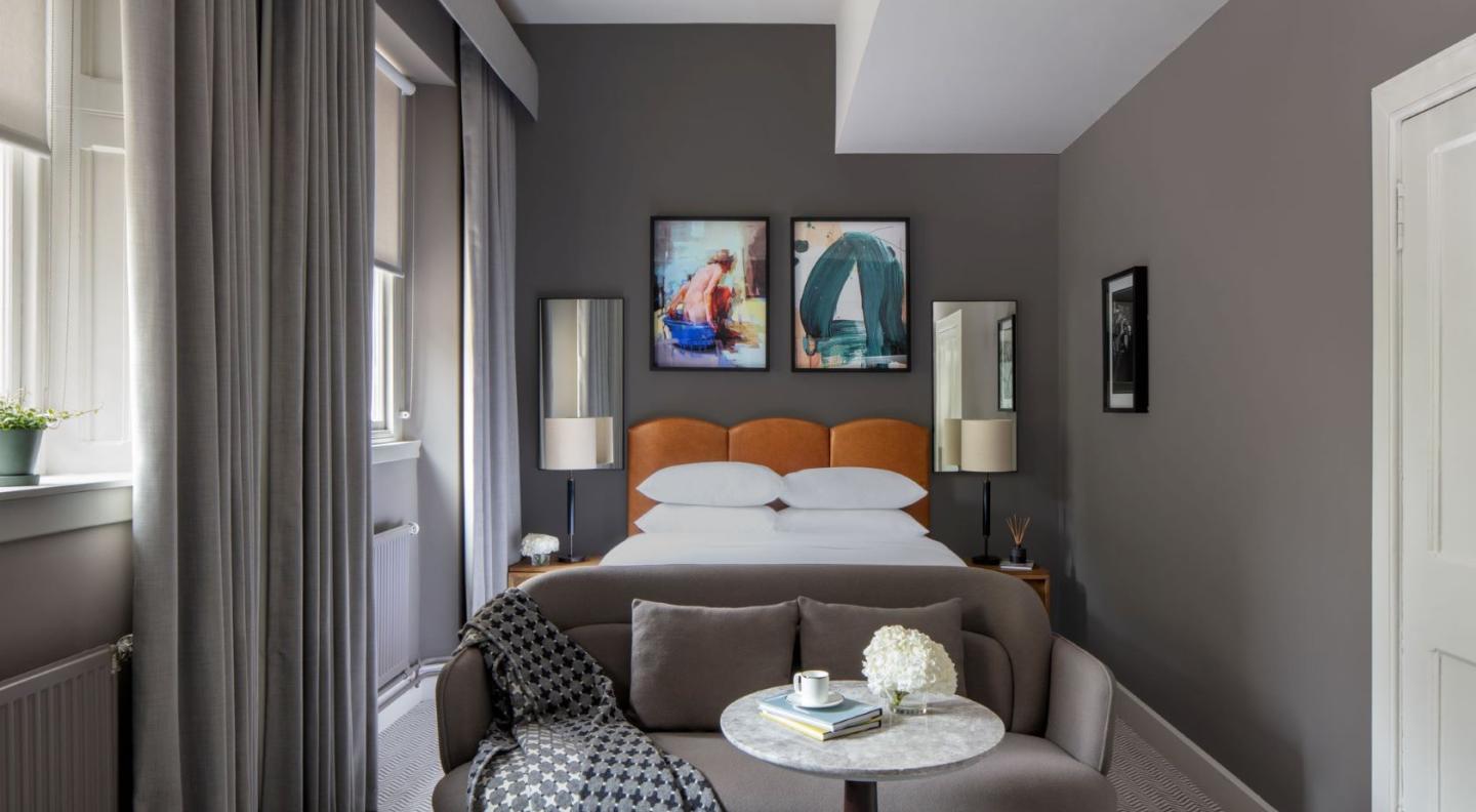 A luxurious hotel bedroom with grey decor, a large bed, colourful paintings, and assorted furrniture, soft furnishings and lamps