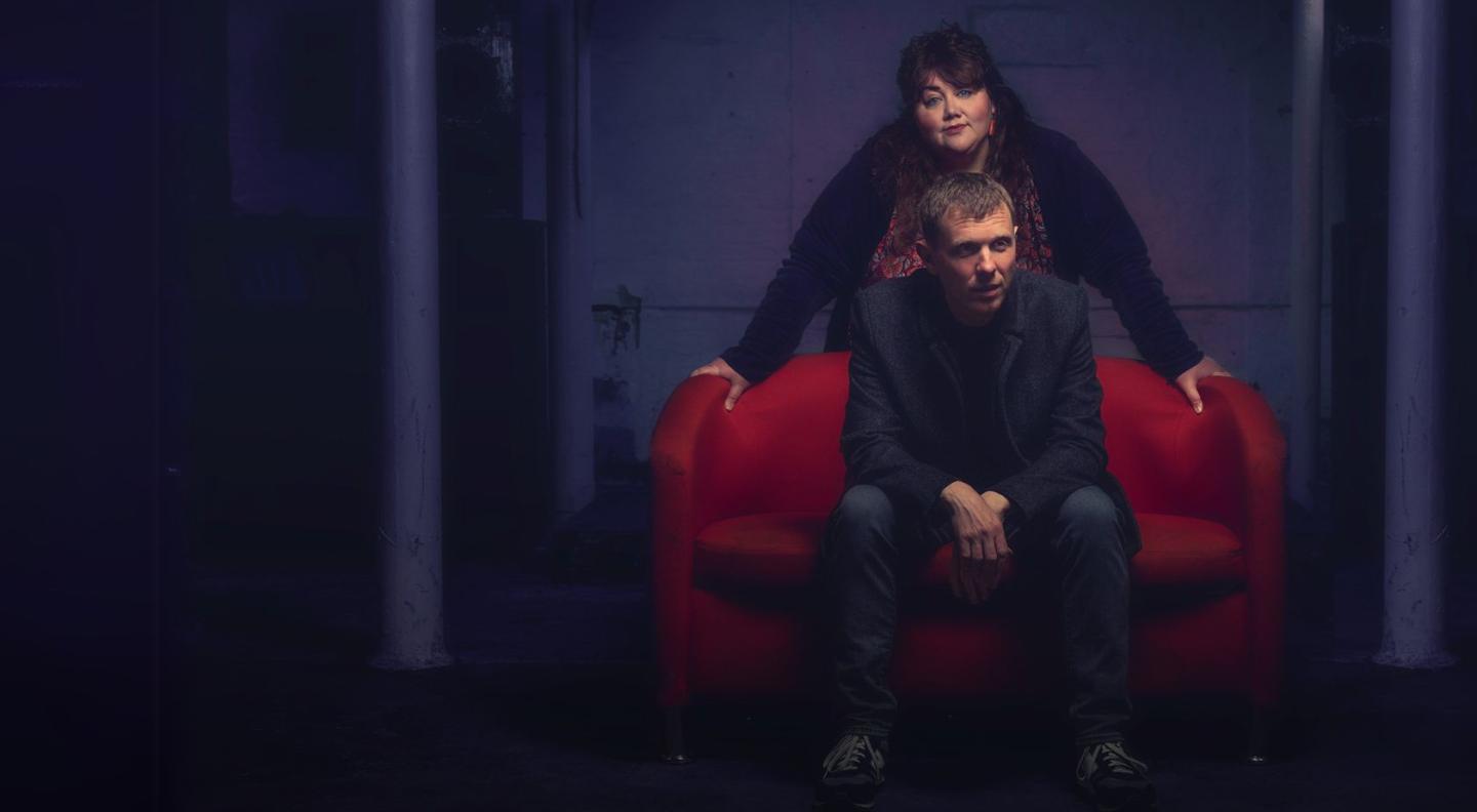 Musicians The Breath - a man with short hair sitting on a red couch, and a woman in black with long black hair standing behind him. 