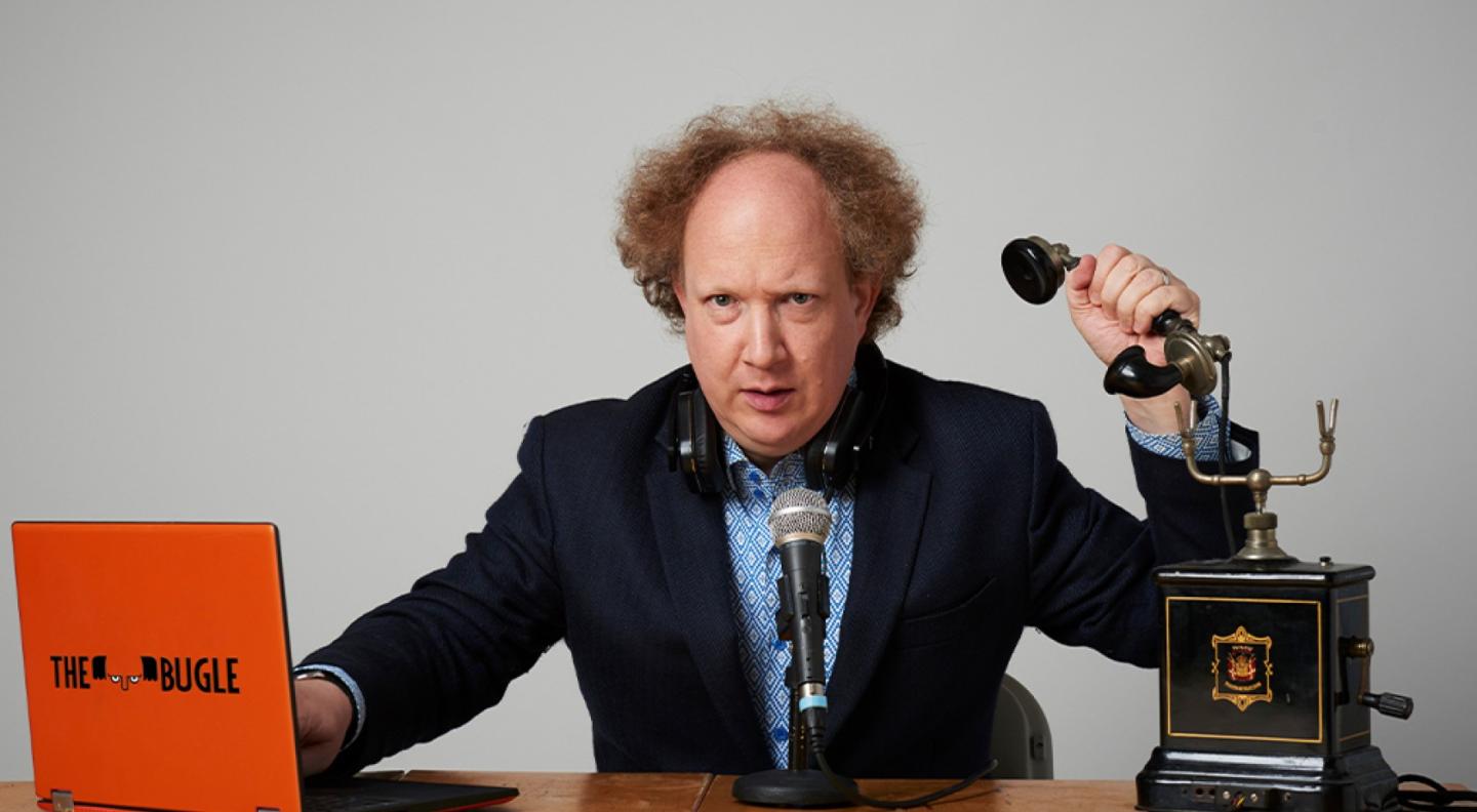 A white man with curly red hair sits behind a desk, holding the receiver of a rotary phone and with an open laptop with The Bugle on it