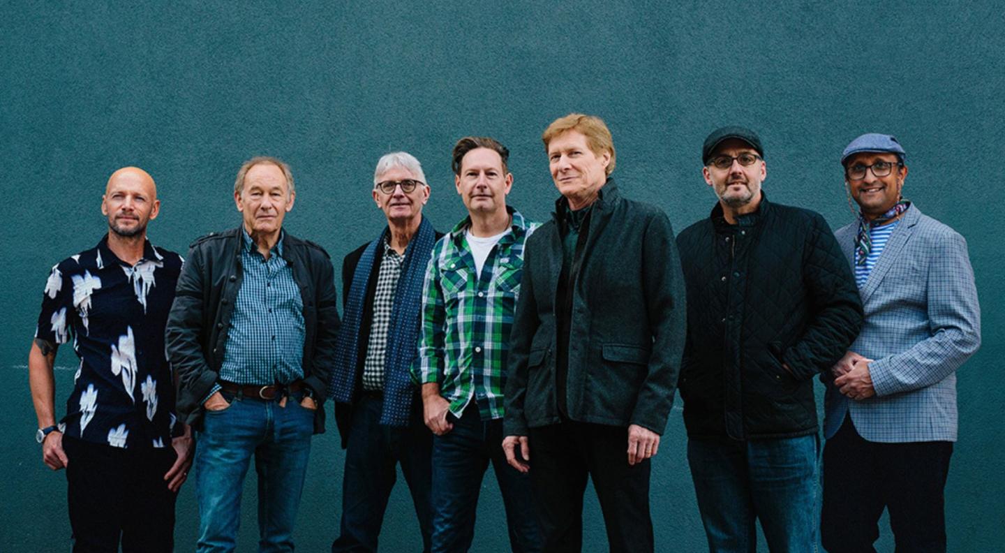The seven members of the band The Manfreds agains a teal blue wall