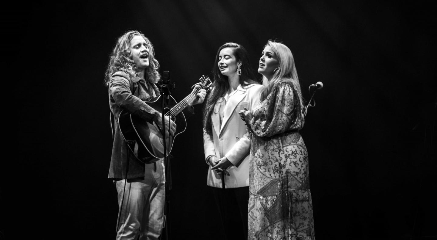A black and white image of the three members of The Wandering Hearts - one white man and two white women - standing in a group singing