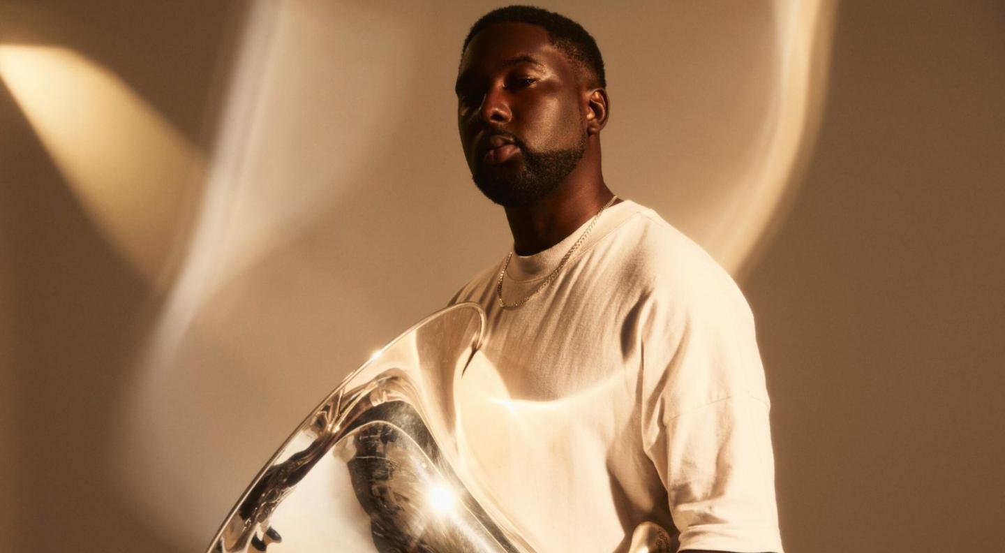 Theon Cross, wearing a white t-shirt, standing in front of a tan background, holding his silver tuba