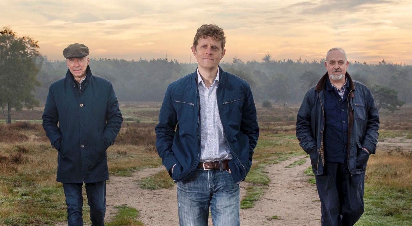 A trio of men wearing outdoor clothing stand in a line, against a background of trees and hills