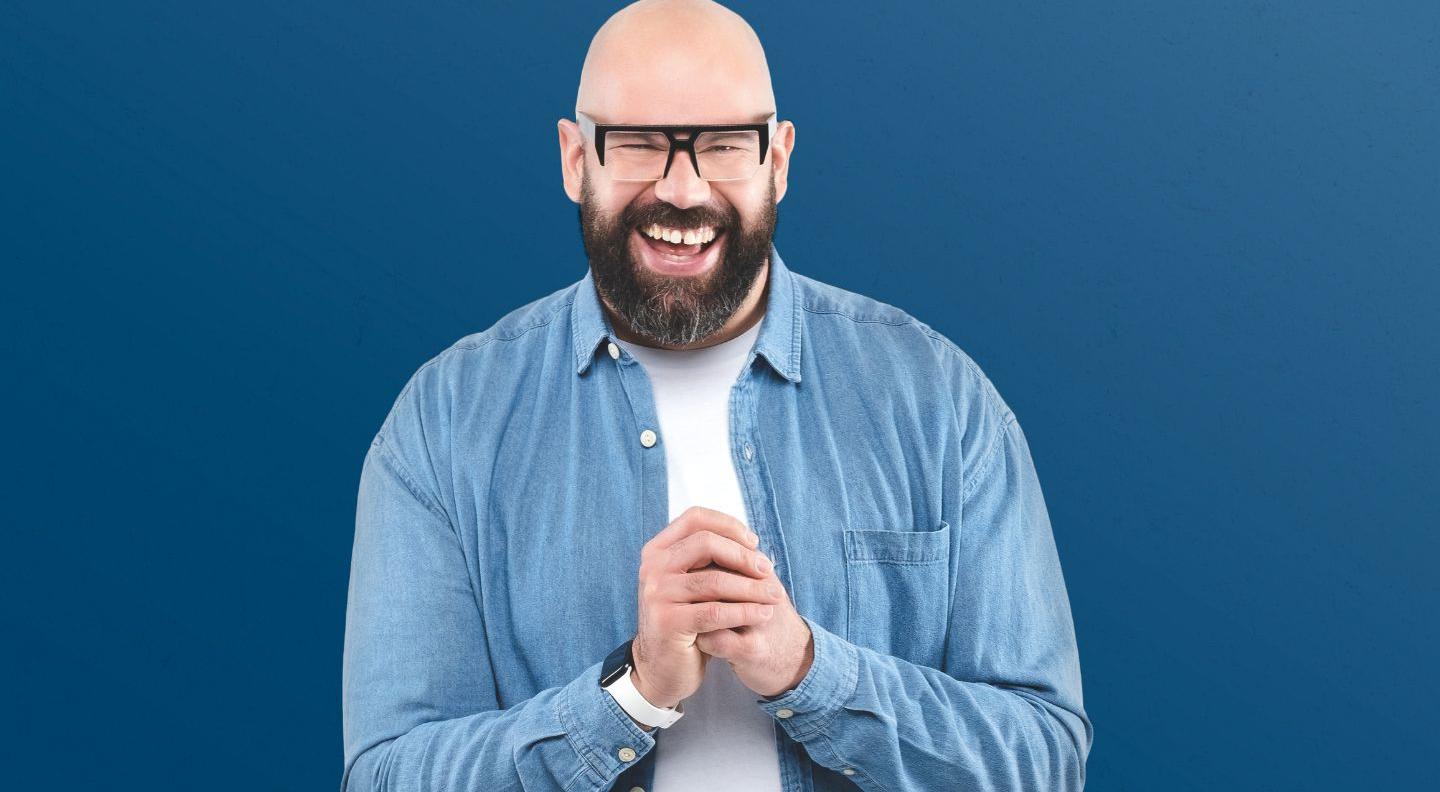 A bald man with a dark beard wears dark-rimmed glasses, standing against a blue background.