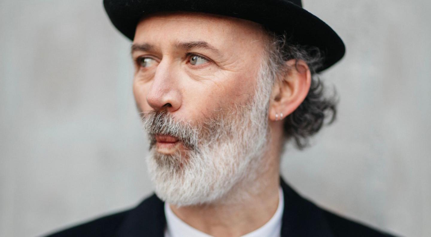 Tommy looks off to his right against a grey background. He's a white man with a greying beard, wearing a clack jacket and hat