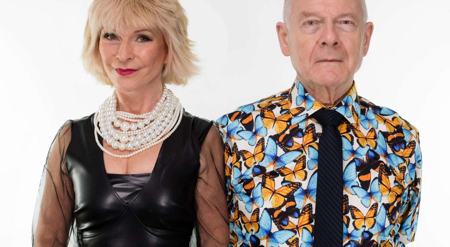 An older white couple face the camera and are shown from the waist up. The woman has chin length peroxide blonde hair and wears red lipstick a large pearl necklace and a black leather dress. The man has receding grey hair and wears a black tie and a long-sleeved shirt with yellow and blue butterflies on it