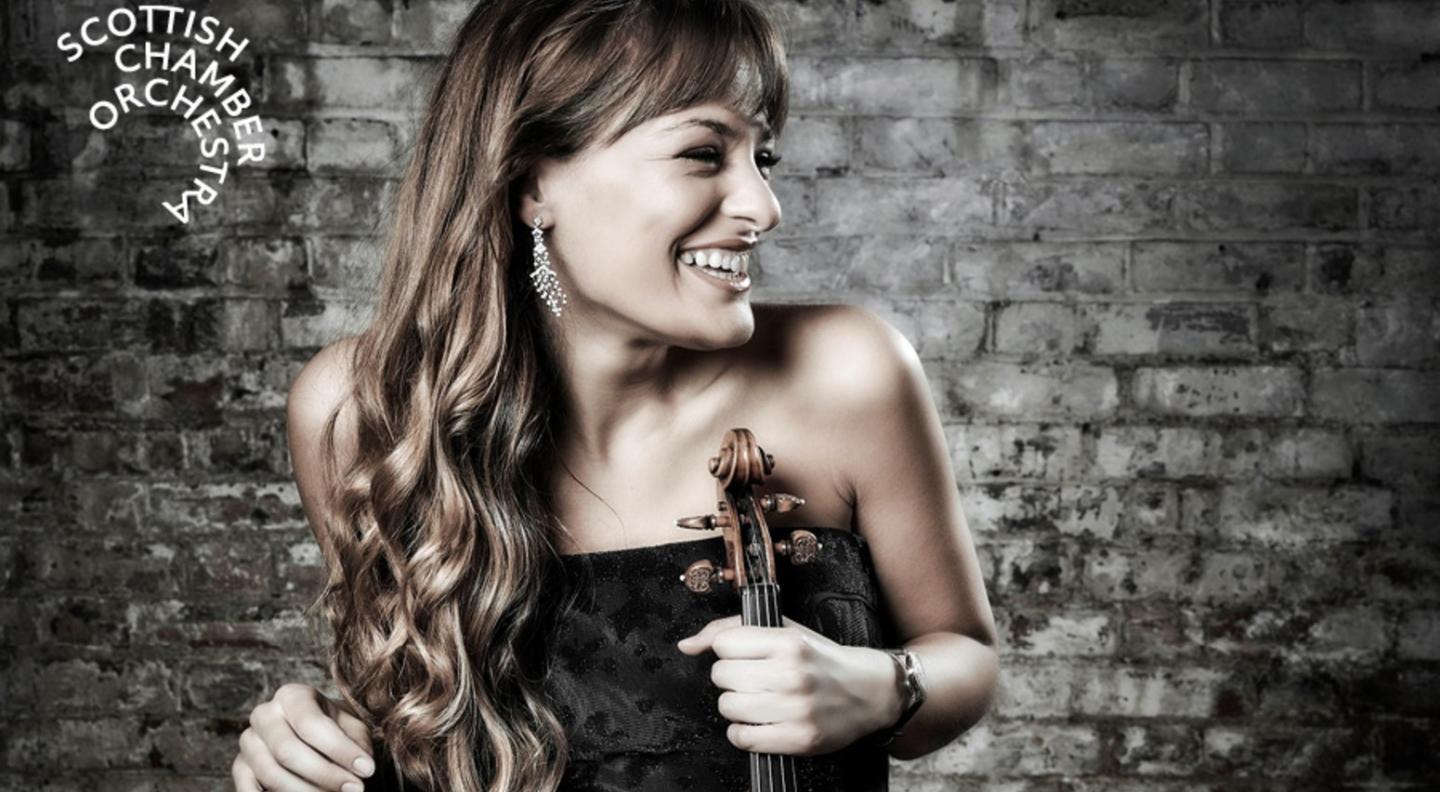 Nicola Benedetti centre frame and smiling holding her violin against a brick wall