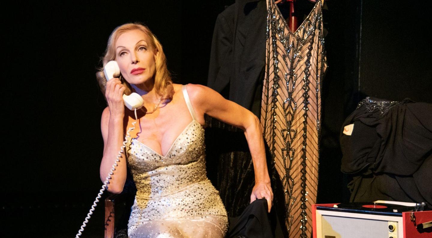 An image of Ute on stage. She wears a sparkling evening gown and holds a rotary phone to her ear. She has blonde hair, styled with vintage curls