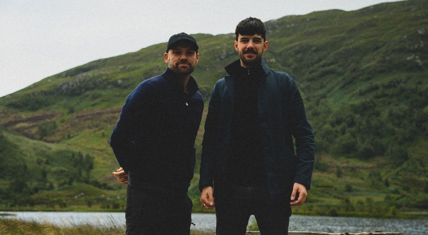 Two white men stand against a background of hills