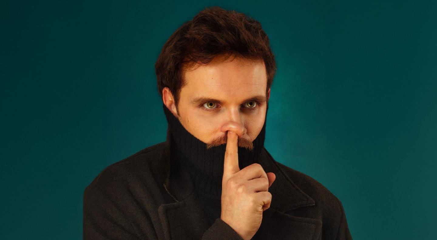Vittorio Angelone in front of a dark blue/green backdrop. He is wearing a black turtleneck jumper, which is pulled up over his chin, and he has one pointer finger up in front of his mouth, in a shh gesture.