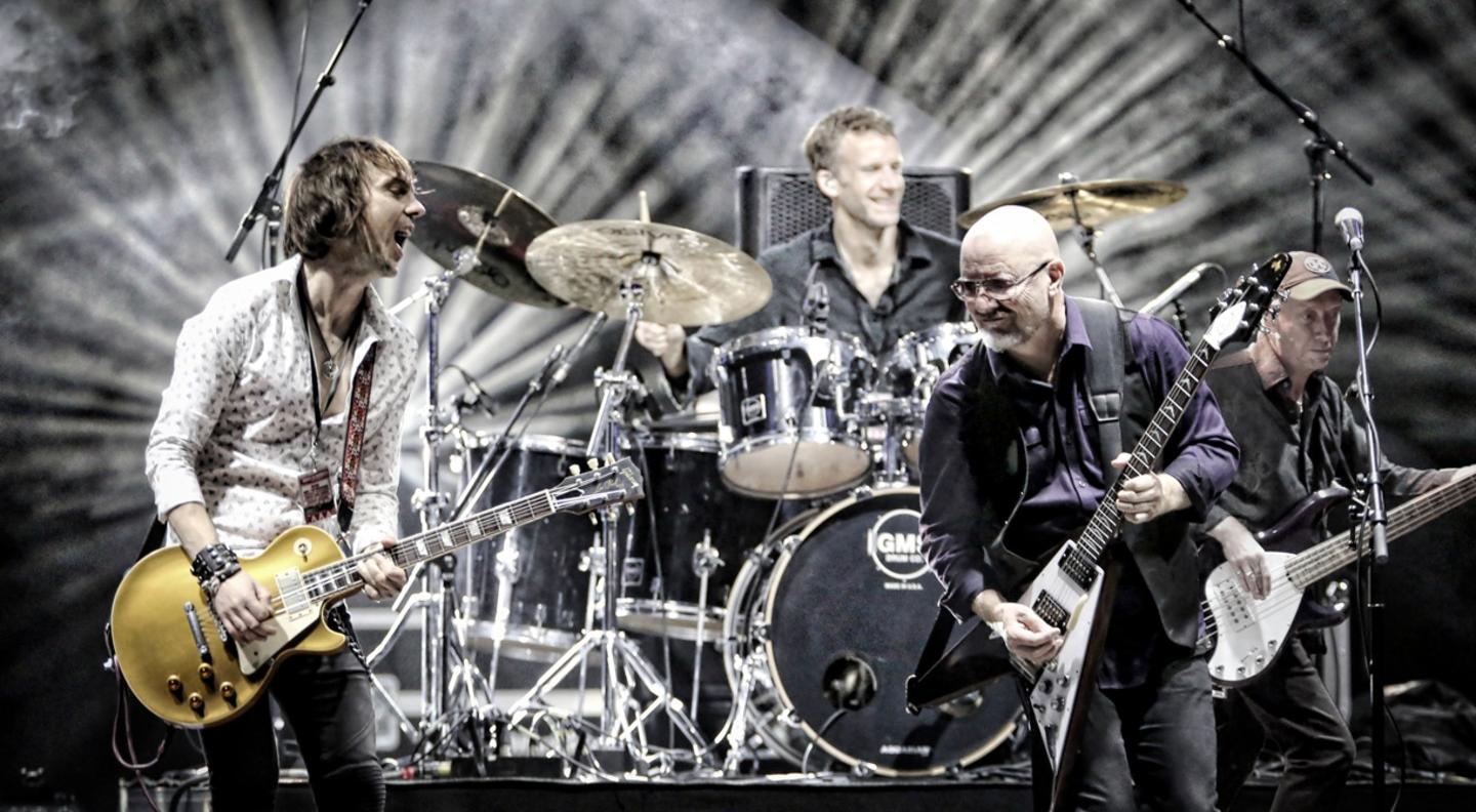 Wishbone Ash are performing on stage with bright lights arcing out behind them