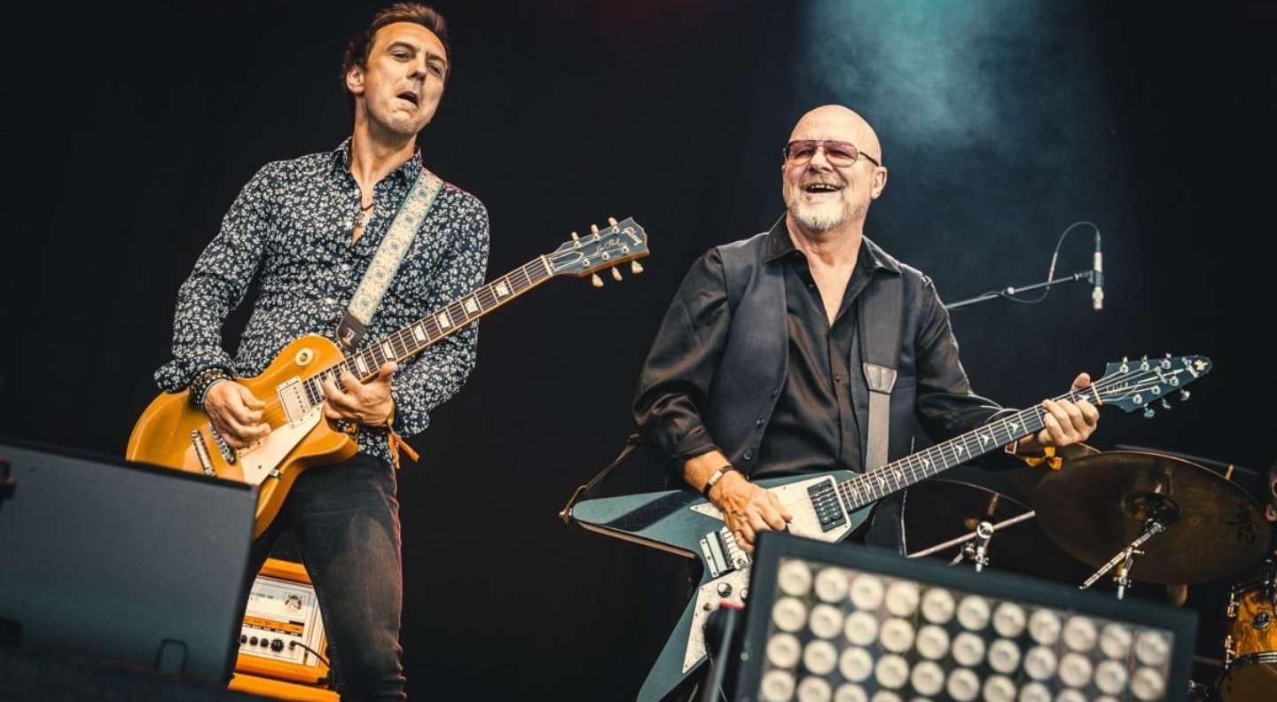 Guitarists Mark Abrahams and Andy Powell of Wishbone Ash play guitar on a large stage, surrounded by speakers and stage lighting