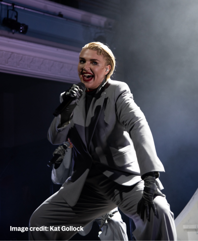 Rebecca Lucy Taylor, or Self Esteem, a white woman in her thirties with short blond hair wears a grey trouser suit and dark gloves. She sings into a microphone and dances