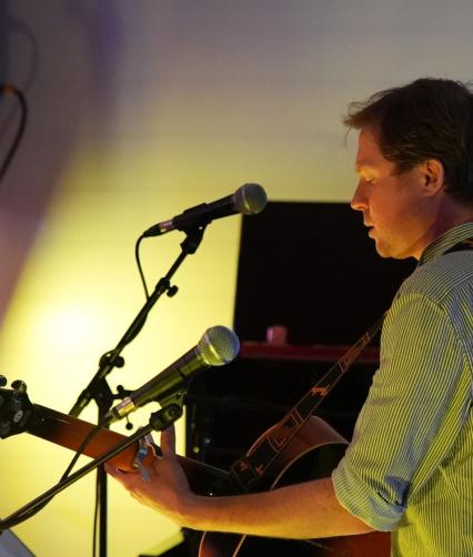 A white man with blonde hair is seen side on standing at a microphone playing guitar