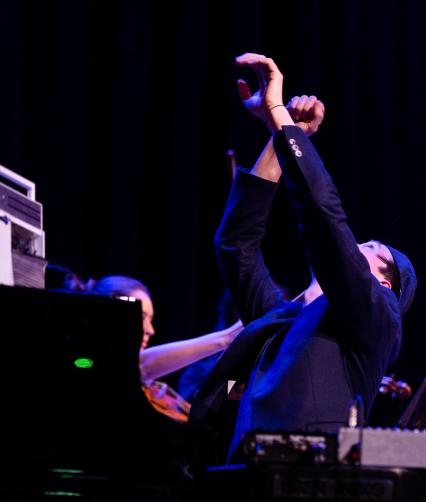 A white man sitting at a piano leans back and crosses his hands in the air