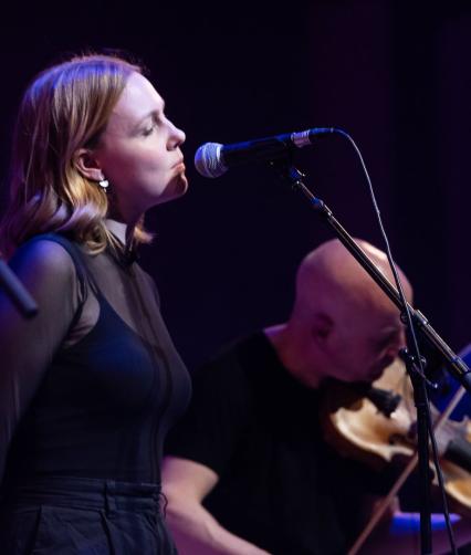 A young white woman with shoulder length fair hair sings into a microphone. A white man behind her plays the fiddle