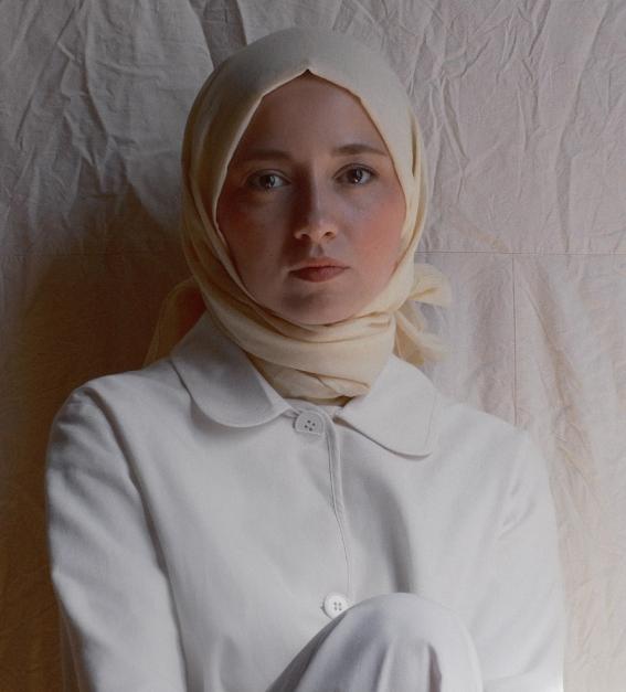 A young woman dressed all in white and wearing a light coloured headscarf leans against a light background with her knee slightly raised looking out at the viewer