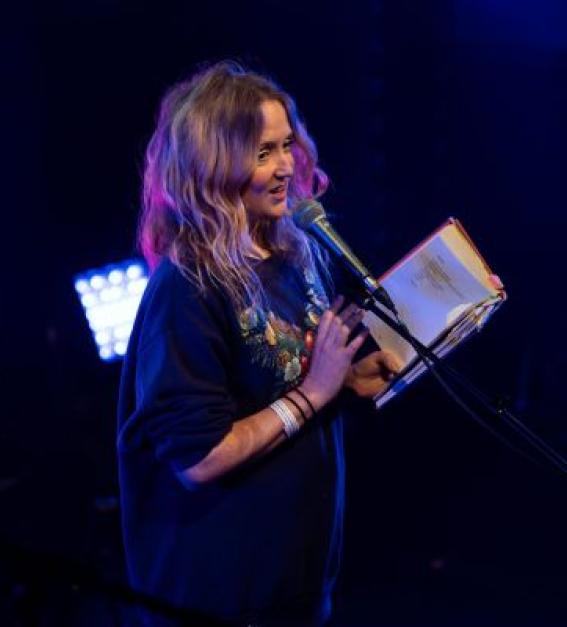 A white woman with long blonde hair is on stage, speaking into a microphone. She has an annotated book of poetry in her hand.