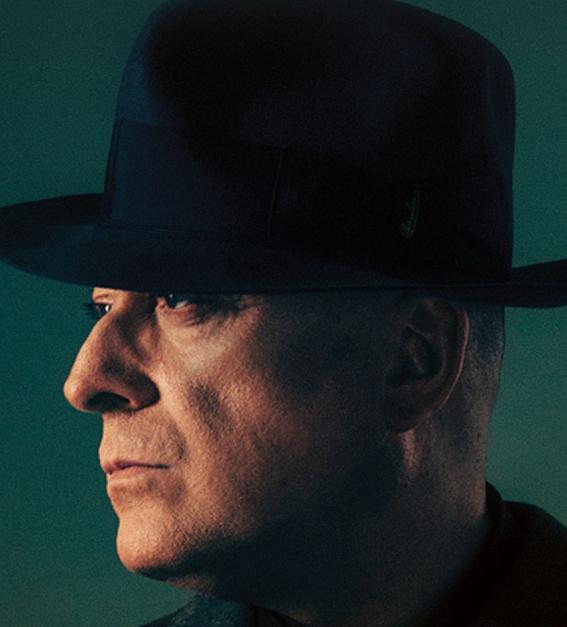 A man in profile, wearing a black bowler hat, against a dark green background 