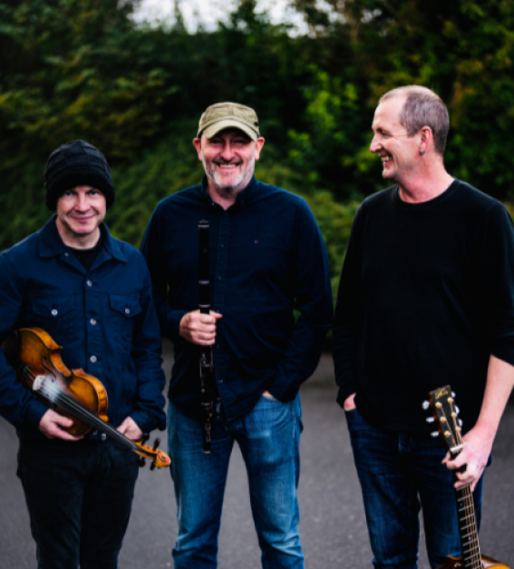 three white middle-aged men holding folk instruments with dark green bushes in the background
