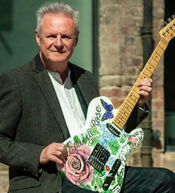 Mike McGeary - a white, middle-aged man with white hair, holds a green guitar in front of a building