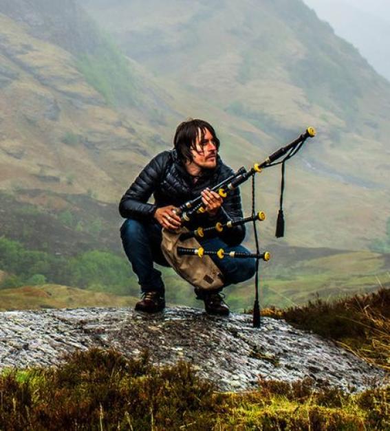 Ross Ainslie with pipes crouching against a beautiful highland backdrop