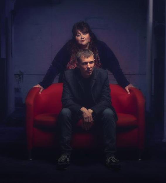 Musicians The Breath - a man with short hair sitting on a red couch, and a woman in black with long black hair standing behind him. 